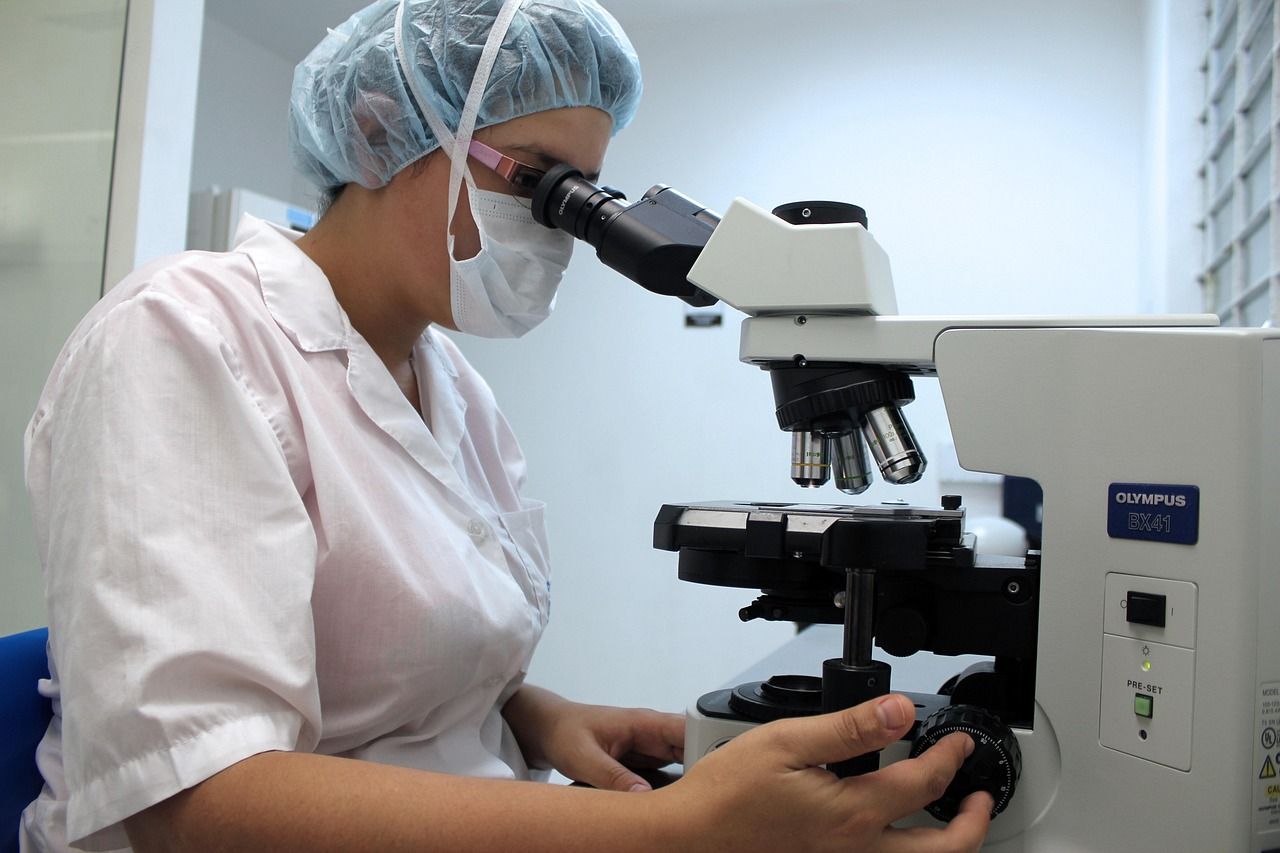 A researcher in a lab coat and hair cover looking through a microscope
