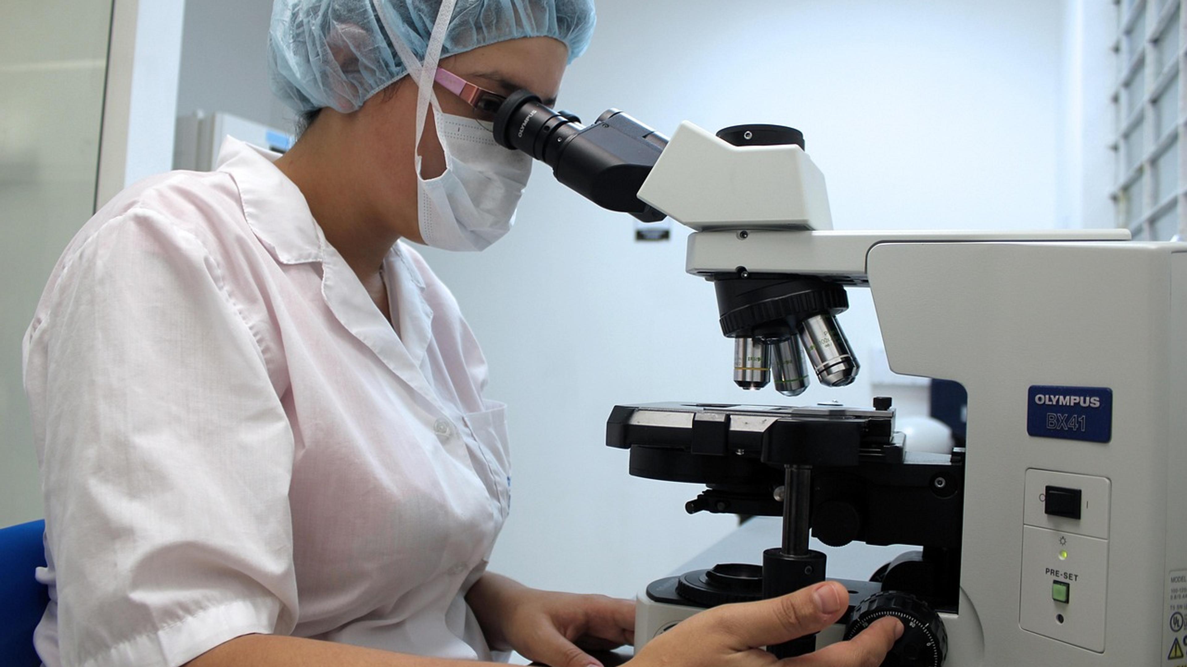 A researcher in a lab coat and hair cover looking through a microscope
