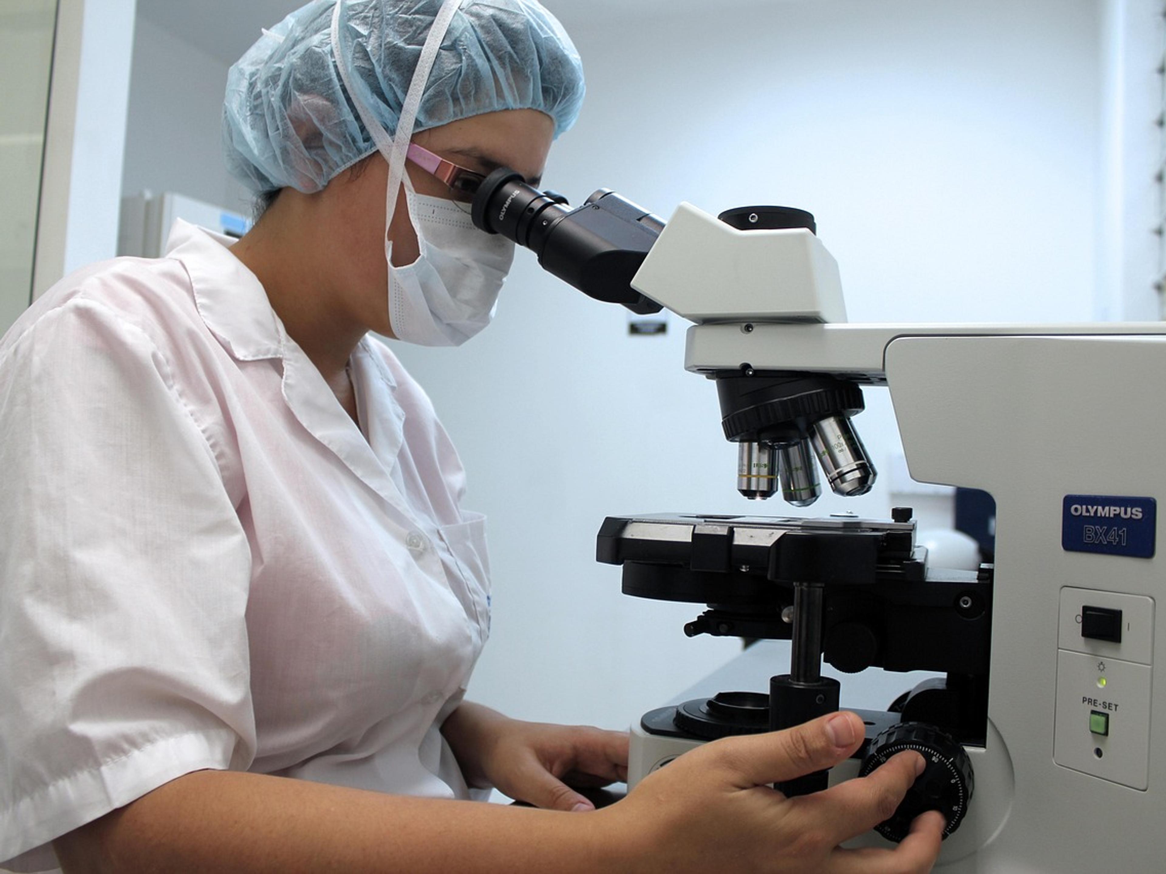 A researcher in a lab coat and hair cover looking through a microscope