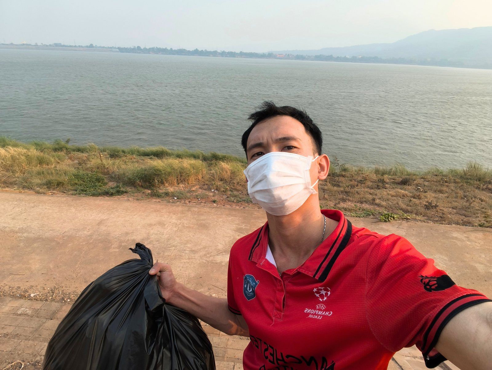 Founder Bounpanh Chaleunsin, known as Panh, in a face mask and red shirt, holding a black trash bag on the Mekong riverbank