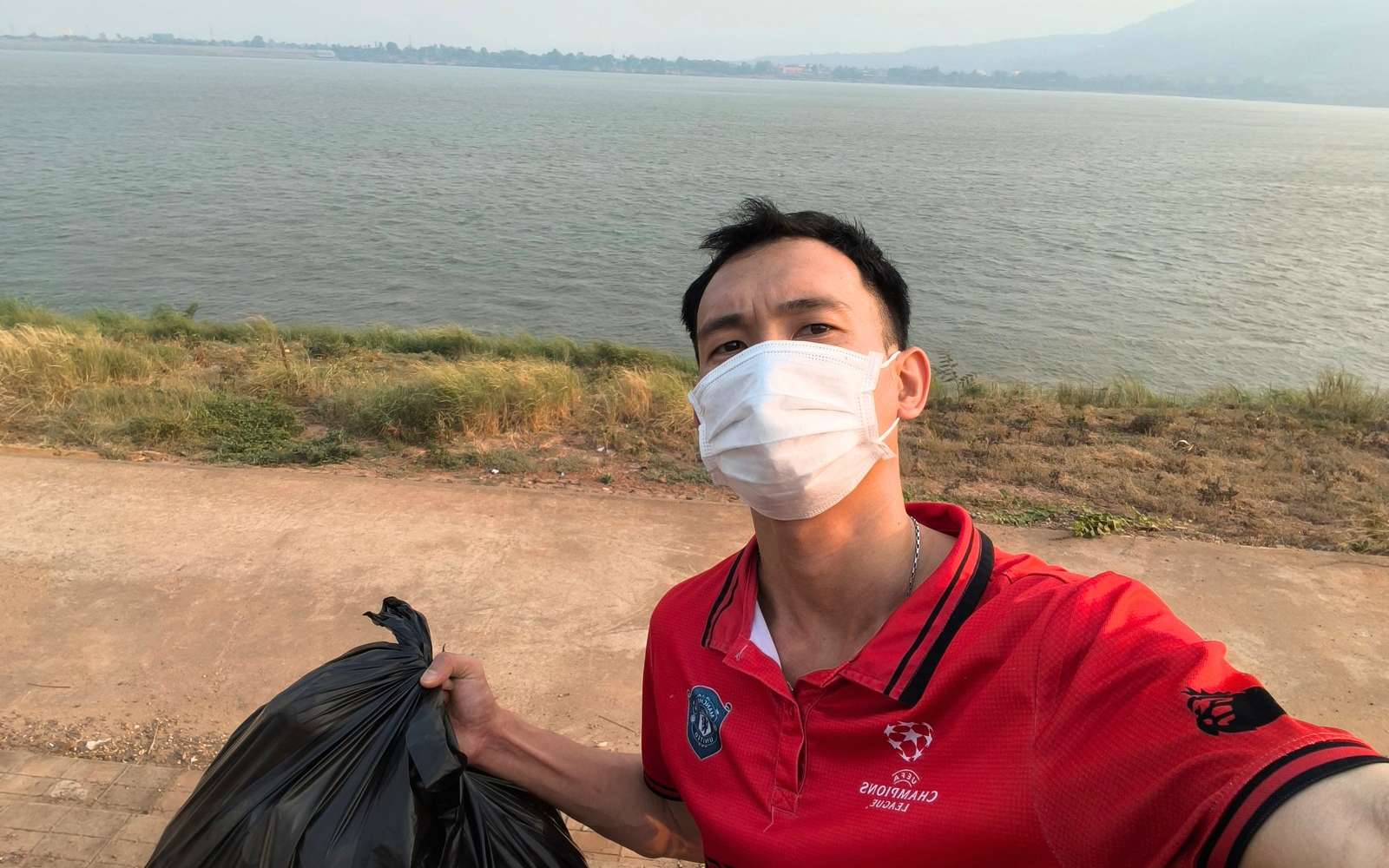 Founder Bounpanh Chaleunsin, known as Panh, in a face mask and red shirt, holding a black trash bag on the Mekong riverbank