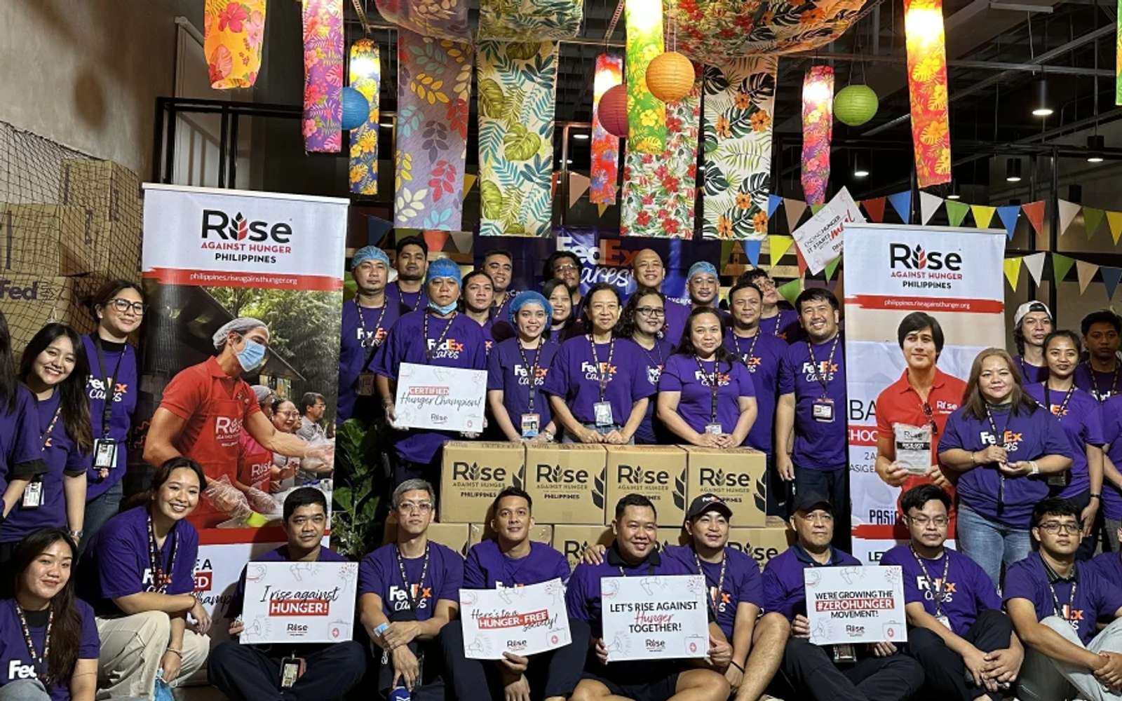 Group photo of FedEx volunteers in purple shirts with Rise Against Hunger Philippines banners, boxed meal kits and hand-written signs