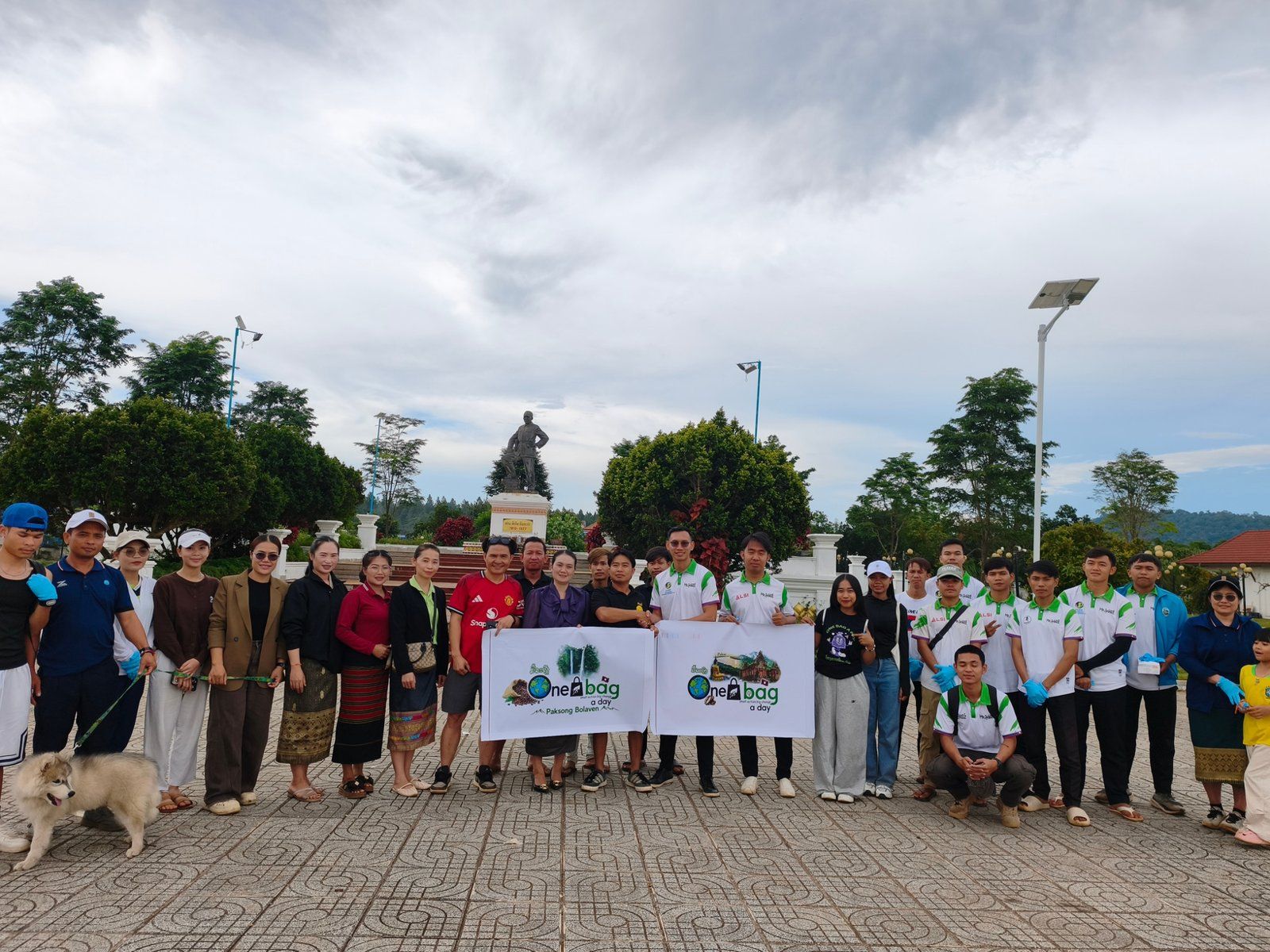 Volunteers holding One Bag A Day banners, with a dog in front, on a paved square with trees and a statue behind them