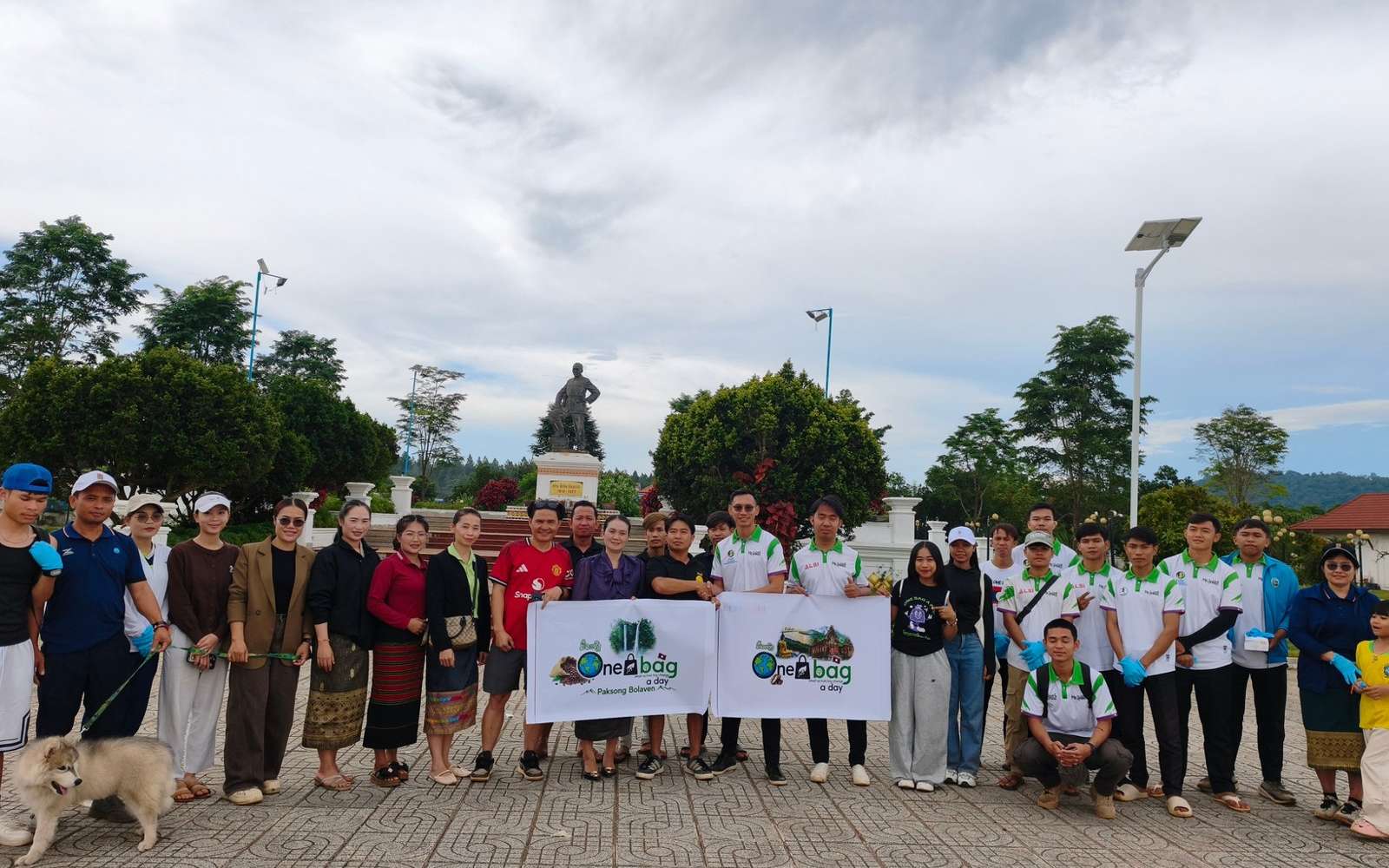 Volunteers holding One Bag A Day banners, with a dog in front, on a paved square with trees and a statue behind them
