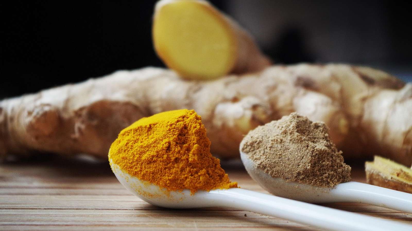 Fresh turmeric and ginger roots beside a spoon of orange turmeric powder on a wooden board