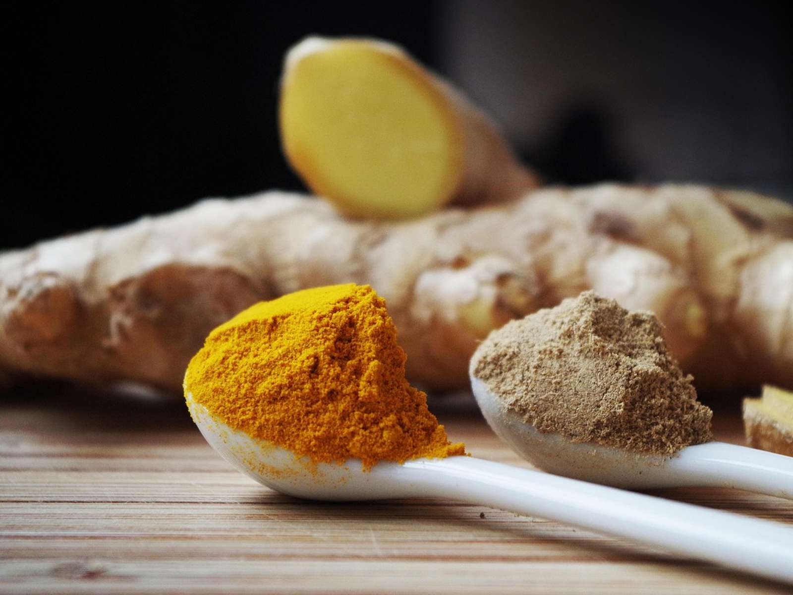 Fresh turmeric and ginger roots beside a spoon of orange turmeric powder on a wooden board