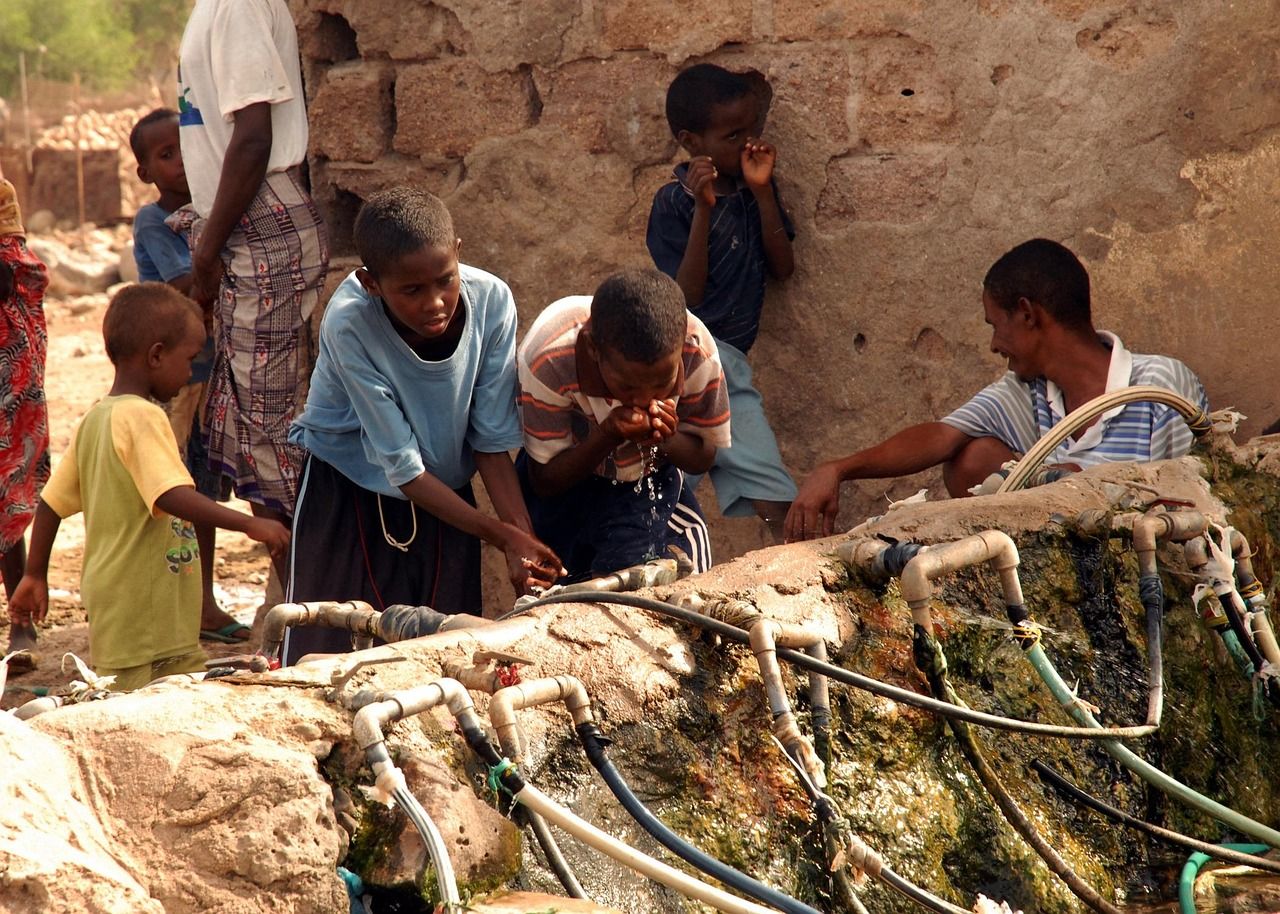 Children and a man drinking from outdoor taps fed by hoses along a stone wall