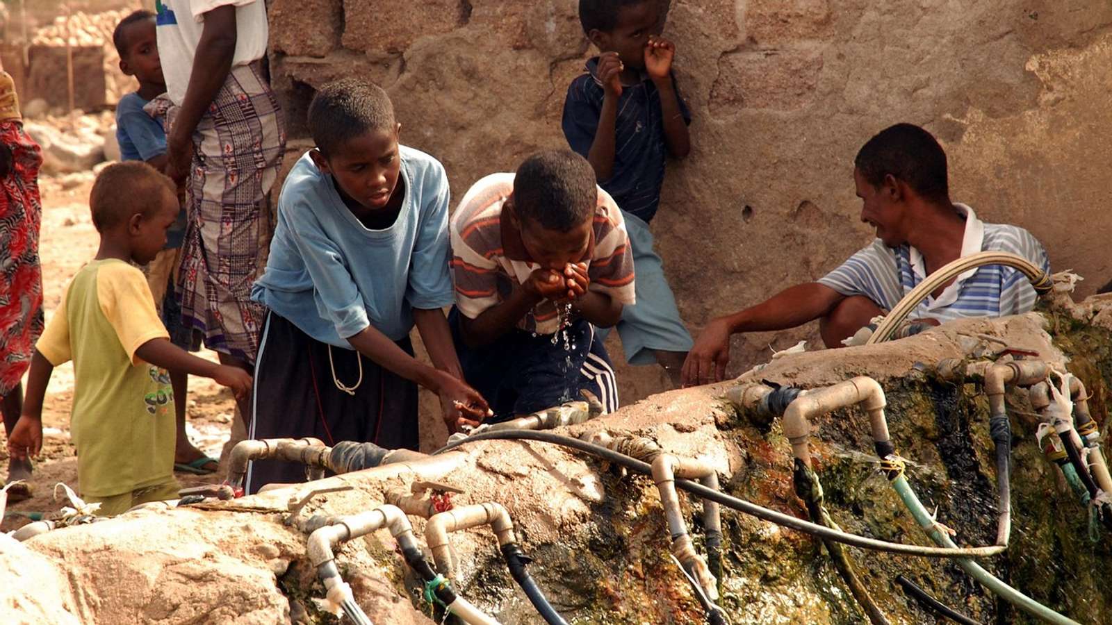 Children and a man drinking from outdoor taps fed by hoses along a stone wall