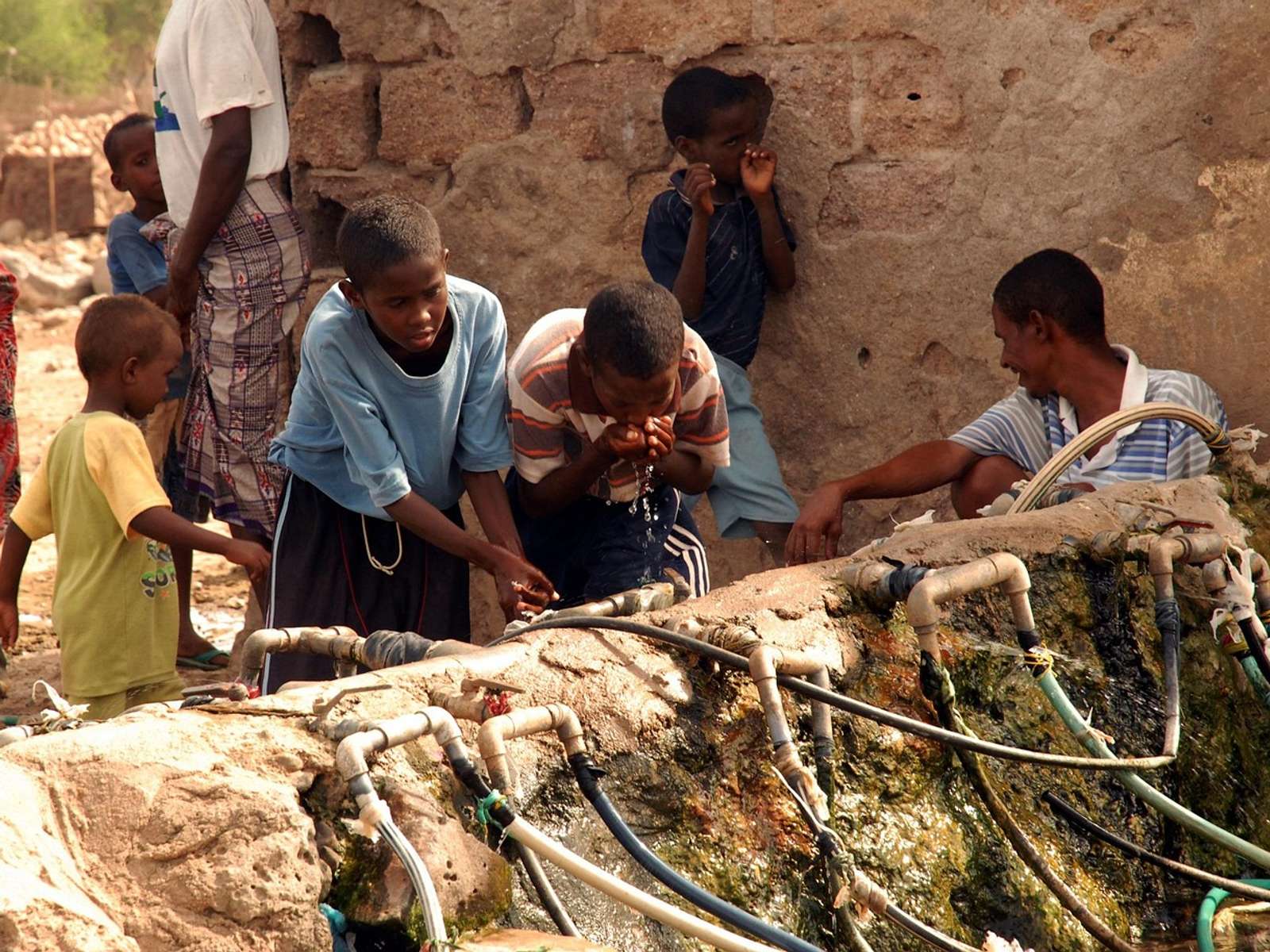 Children and a man drinking from outdoor taps fed by hoses along a stone wall