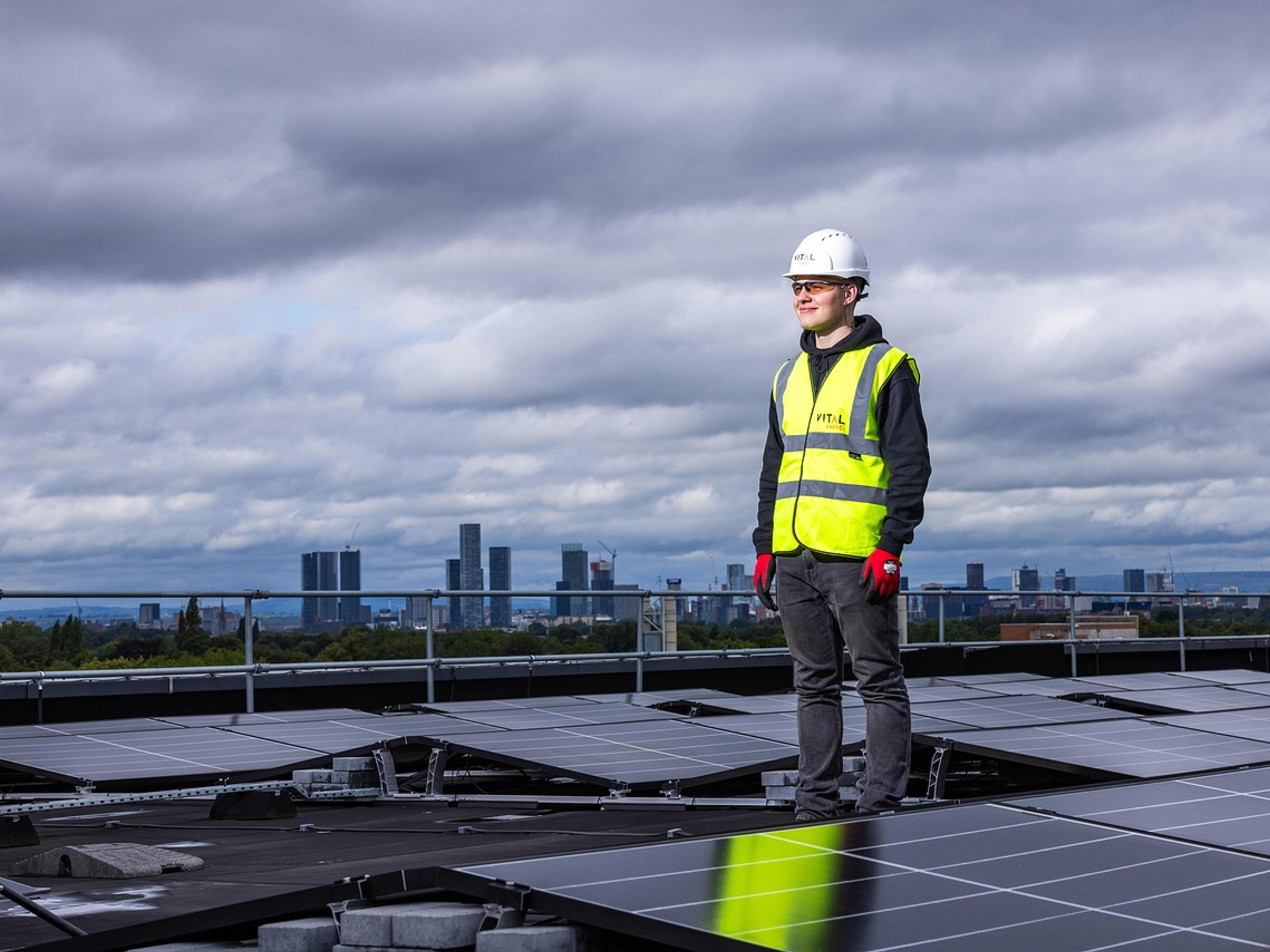 A worker in a hard hat and high-visibility vest standing among rooftop solar panels