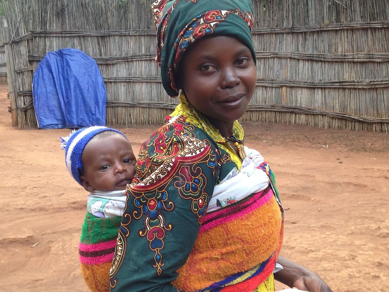 A smiling African woman in a patterned dress carrying a baby on her back
