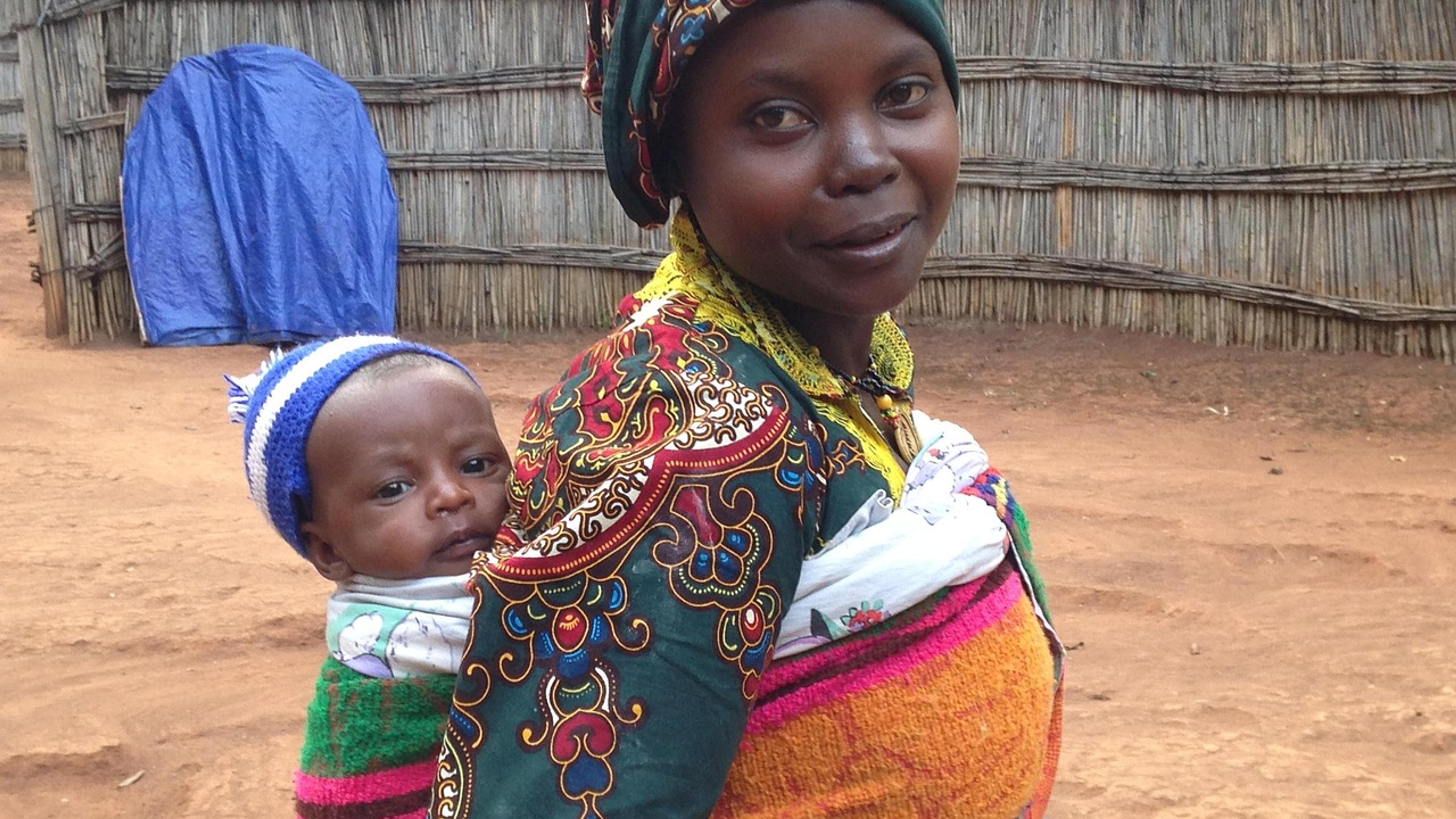 A smiling African woman in a patterned dress carrying a baby on her back