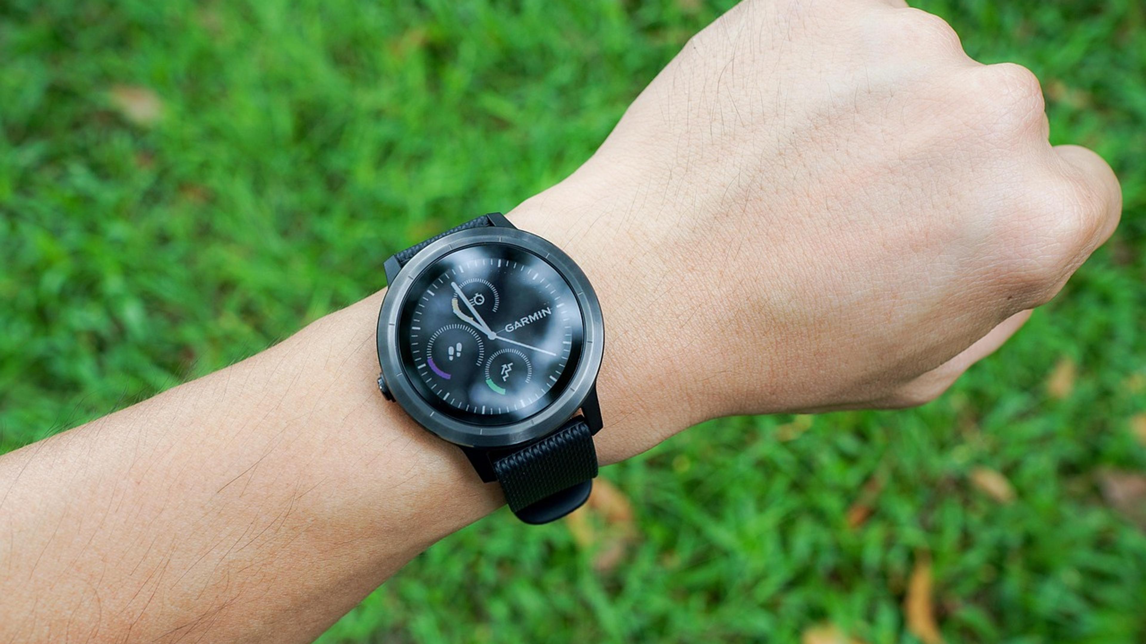A person's wrist wearing a black smartwatch above green grass