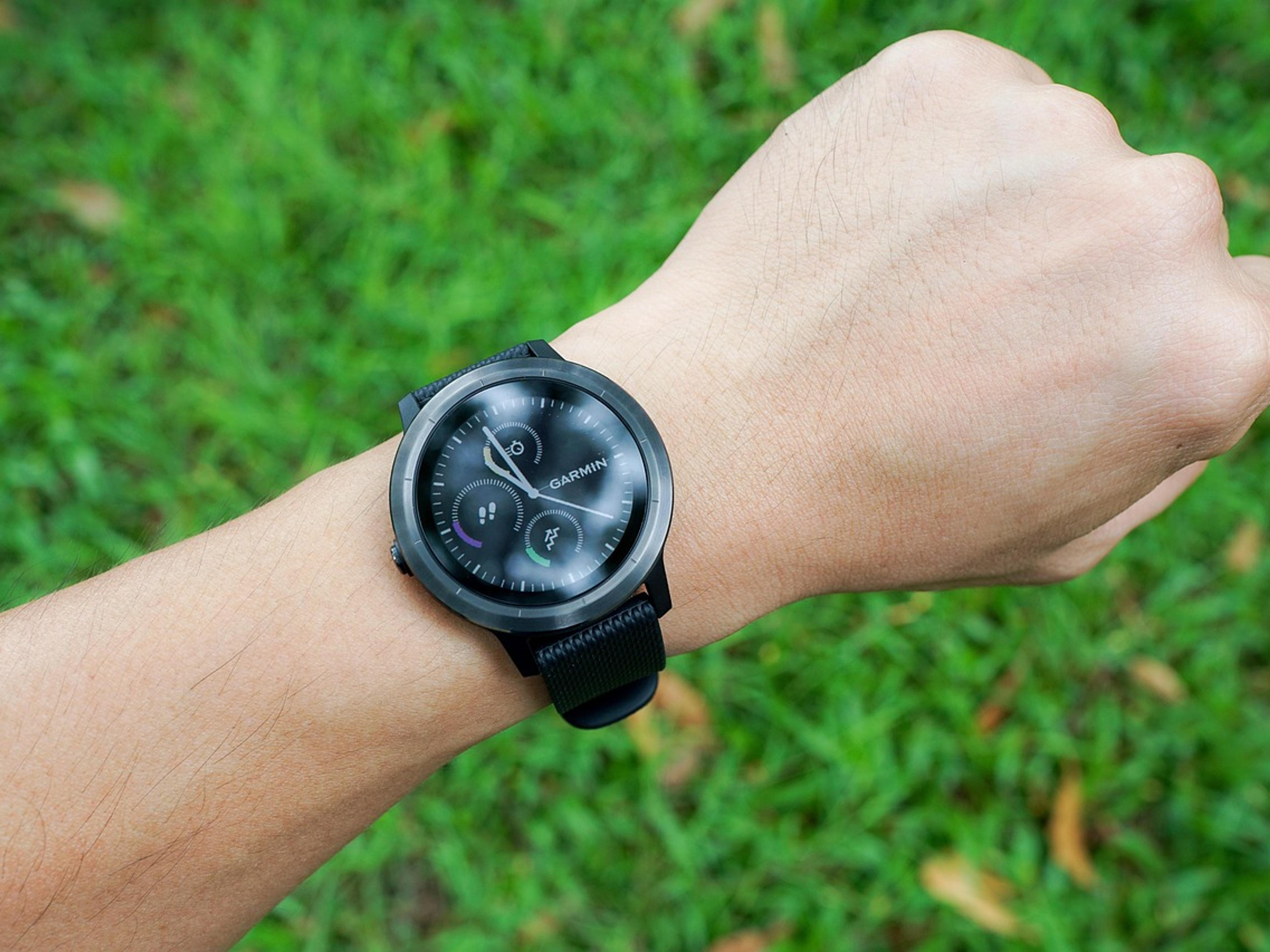 A person's wrist wearing a black smartwatch above green grass