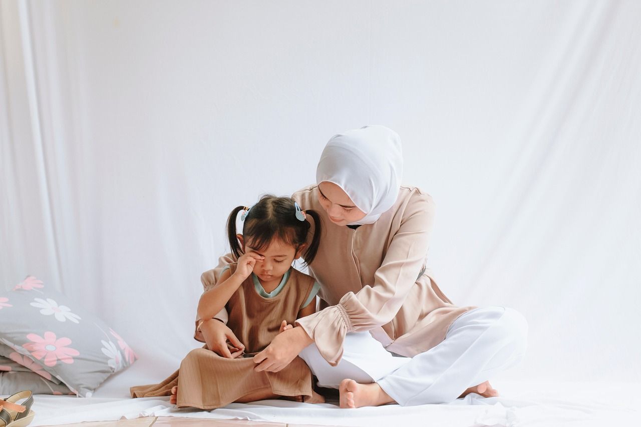 A mother in a headscarf sitting on the floor with her young daughter leaning against her