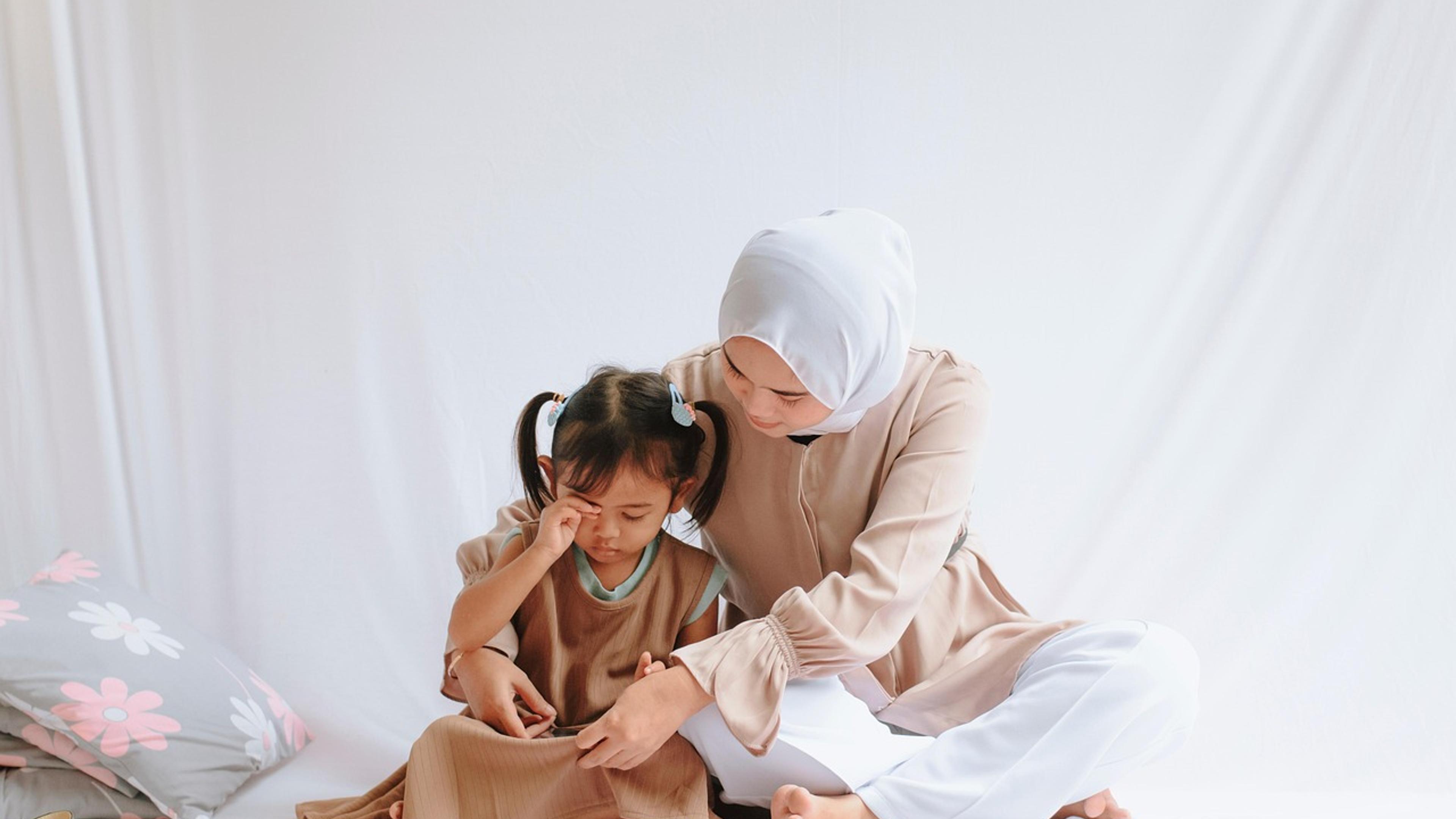 A mother in a headscarf sitting on the floor with her young daughter leaning against her