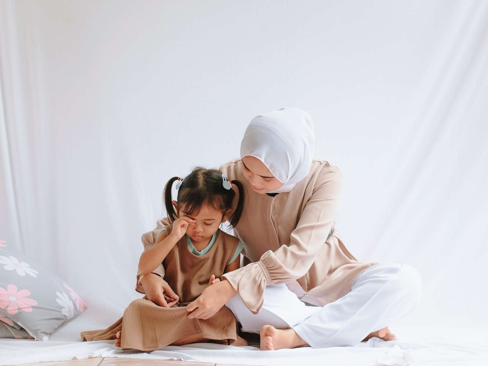 A mother in a headscarf sitting on the floor with her young daughter leaning against her
