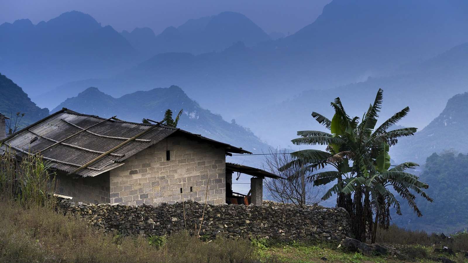A small brick house with a corrugated roof and a banana tree in front of misty mountains in Ha Giang, northern Vietnam