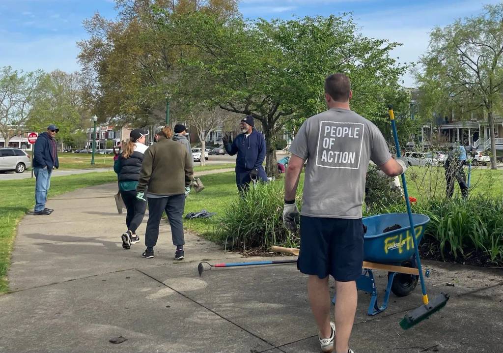 A volunteer wearing a 'People of Action' shirt works in a community garden bed at Chimborazo Park, while other volunteers walk and work in the background on a sunny spring day with flowering trees and residential homes visible.