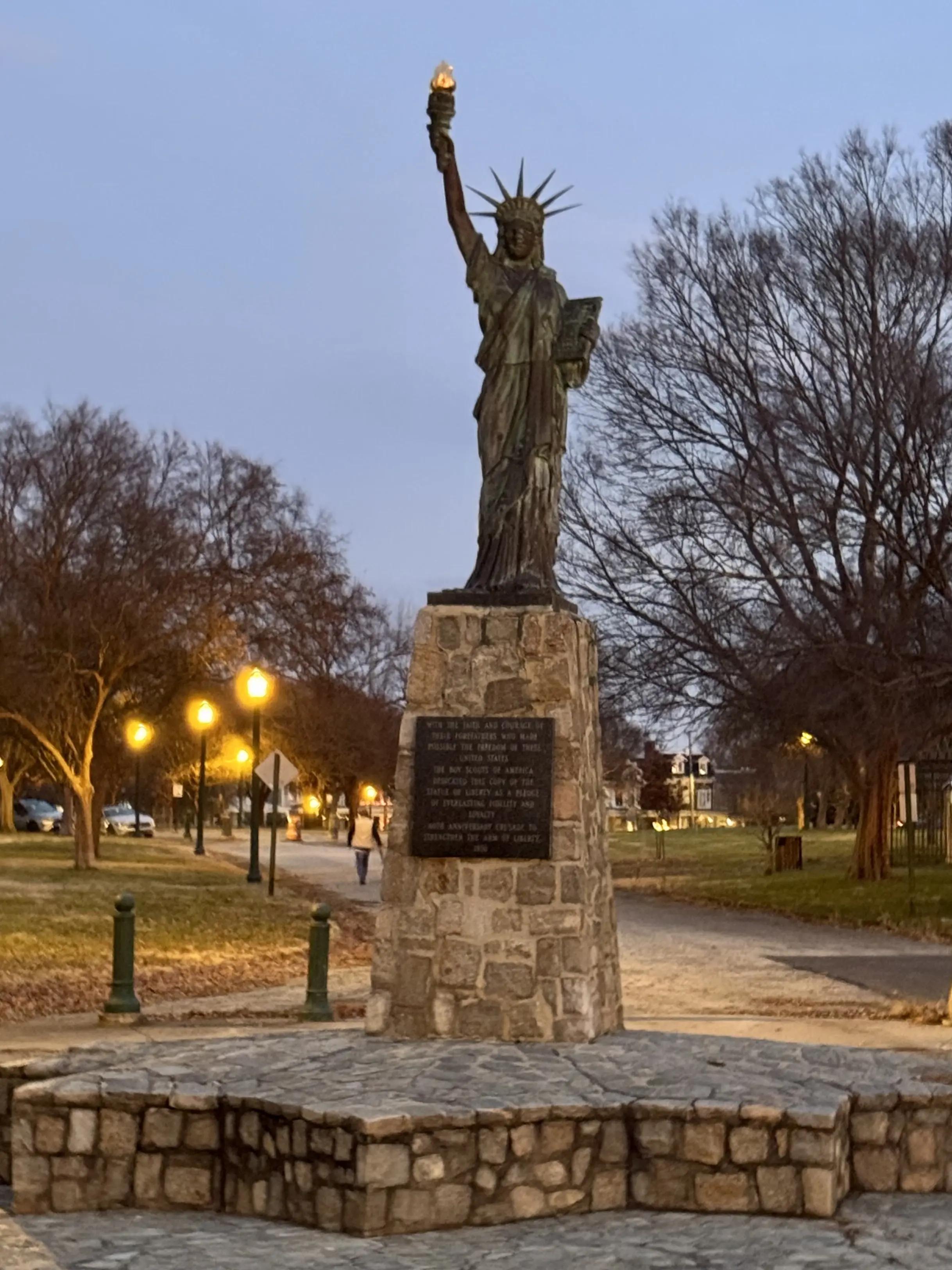 A replica of the Statue of Liberty stands illuminated at twilight in Chimborazo Park, mounted on a stone pedestal with a plaque, surrounded by bare winter trees and lit pathways.