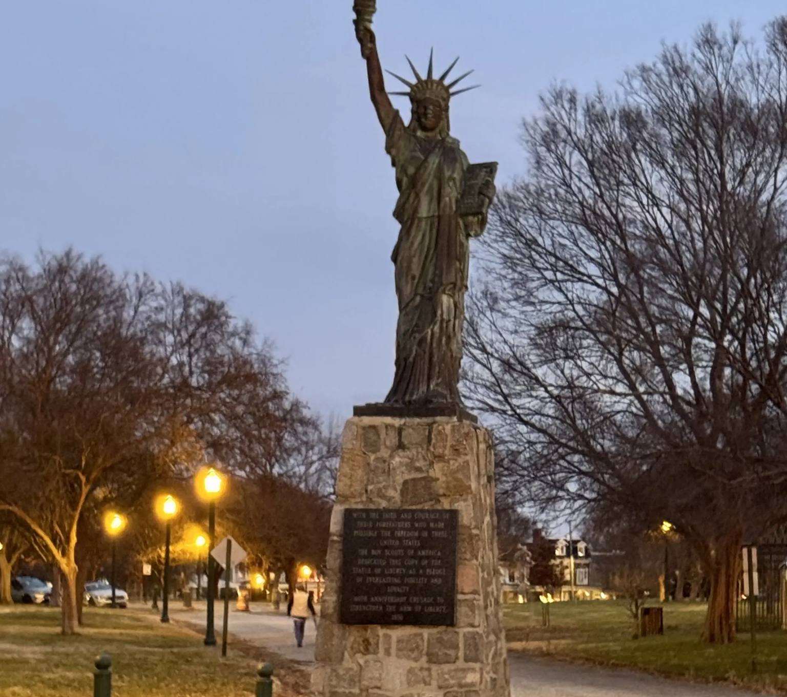 A replica of the Statue of Liberty stands illuminated at twilight in Chimborazo Park, mounted on a stone pedestal with a plaque, surrounded by bare winter trees and lit pathways.