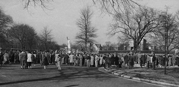 Black and white photograph showing a large crowd of people gathered around a monument in Chimborazo Park, with bare trees lining the background and historic buildings visible in the distance.