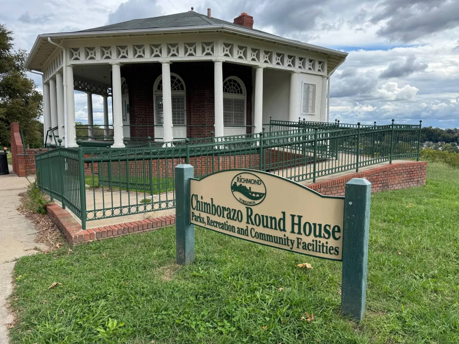 Alt Text "The historic Chimborazo Round House, a Victorian-era pavilion with white Classical columns, decorative frieze, and red brick foundation, surrounded by ornamental green iron fencing. A Richmond Parks and Recreation sign stands in the foreground under dramatic cloudy skies.
