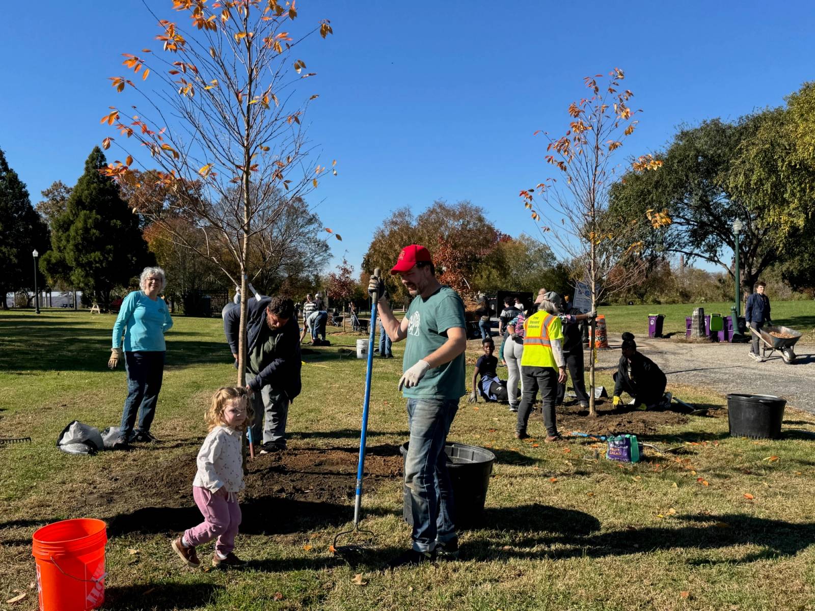 A community tree planting event at Chimborazo Park where volunteers of various ages, including children and adults, plant trees together on a sunny autumn day with fall foliage visible in the background.