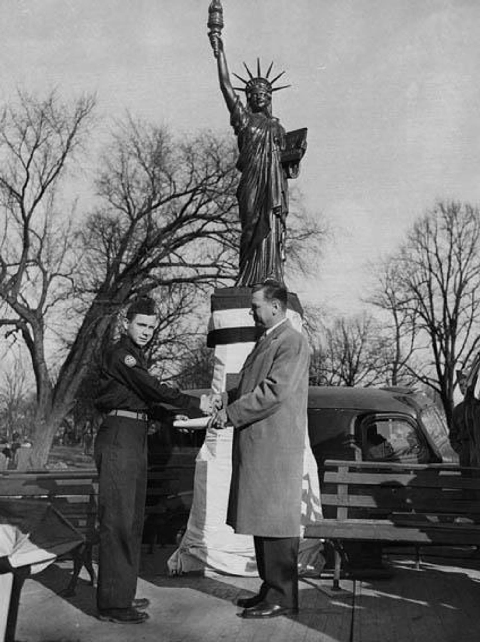 A black and white photograph showing a police officer and a man in a suit exchanging or presenting a document in front of a replica of the Statue of Liberty at Chimborazo Park, with bare trees and vintage cars visible in the background.