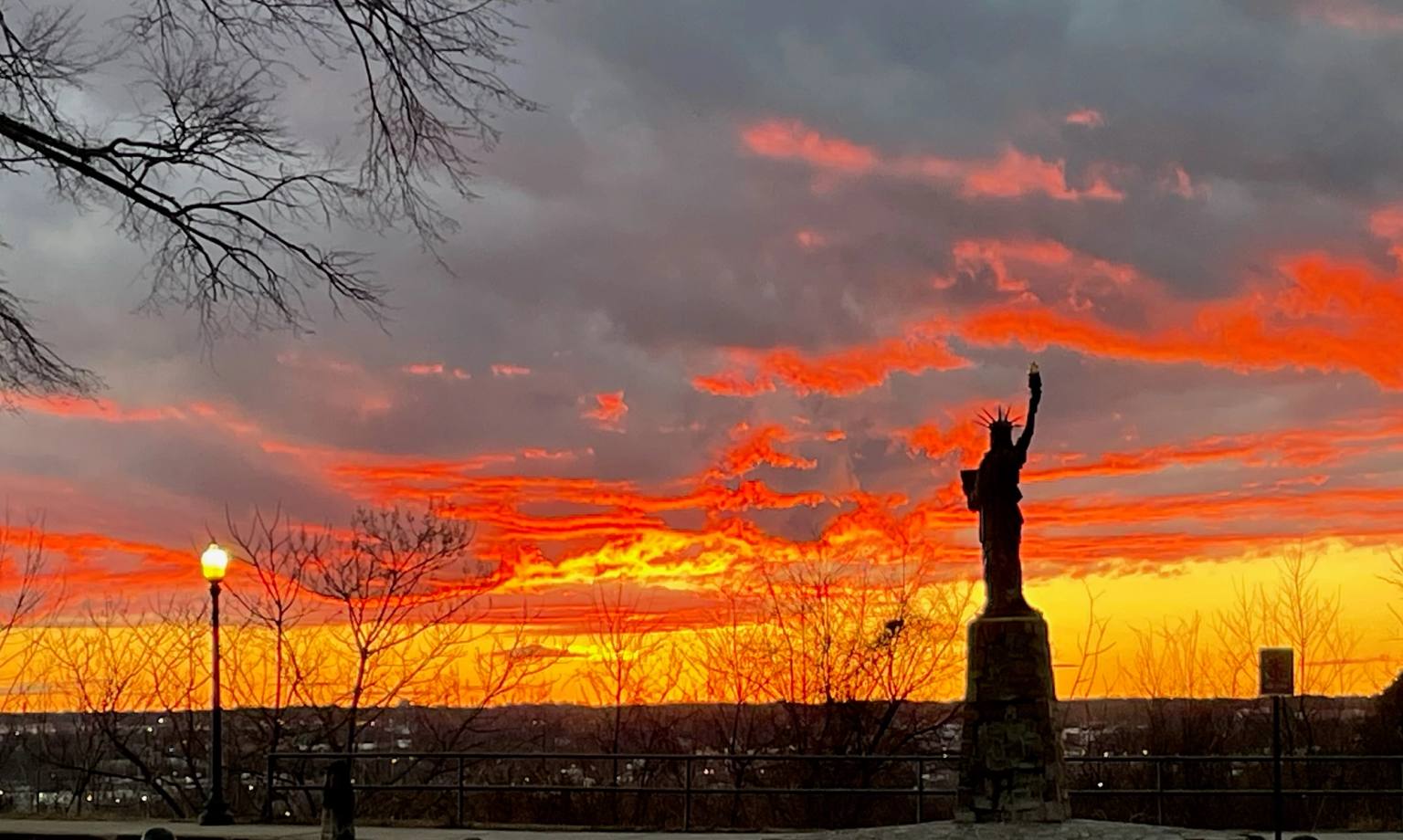 Silhouette of a Statue of Liberty replica monument against a dramatic orange and yellow sunset sky at Chimborazo Park in Richmond, Virginia, with bare winter trees and a street lamp visible in the foreground.