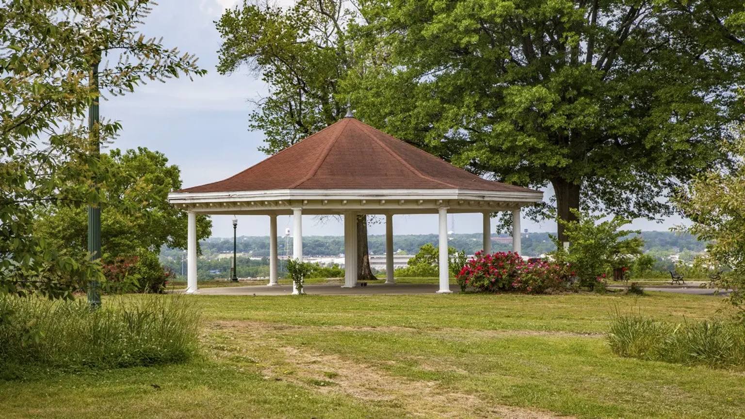 A white octagonal gazebo with a brown roof sits on a grassy lawn in Chimborazo Park, surrounded by mature trees and flowering shrubs, with a distant view of Richmond's landscape and tree-lined hills visible beyond.