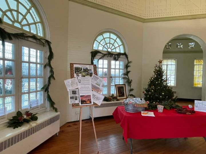 The Chimborazo Park Round House room decorated for the holiday season featuring a fireplace with red poinsettias, a wreath with red ribbon above the mantel, a decorated Christmas tree on the left, and two tables draped in red tablecloths with refreshments and displays.