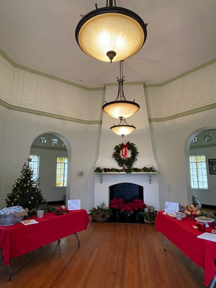 The interior of the Chimborazo Round House with high ceilings features three hanging pendant lights above a white fireplace mantel decorated with a red ribbon wreath and red poinsettias. Red tablecloths drape across tables on either side of the room, with a decorated Christmas tree visible on the left.