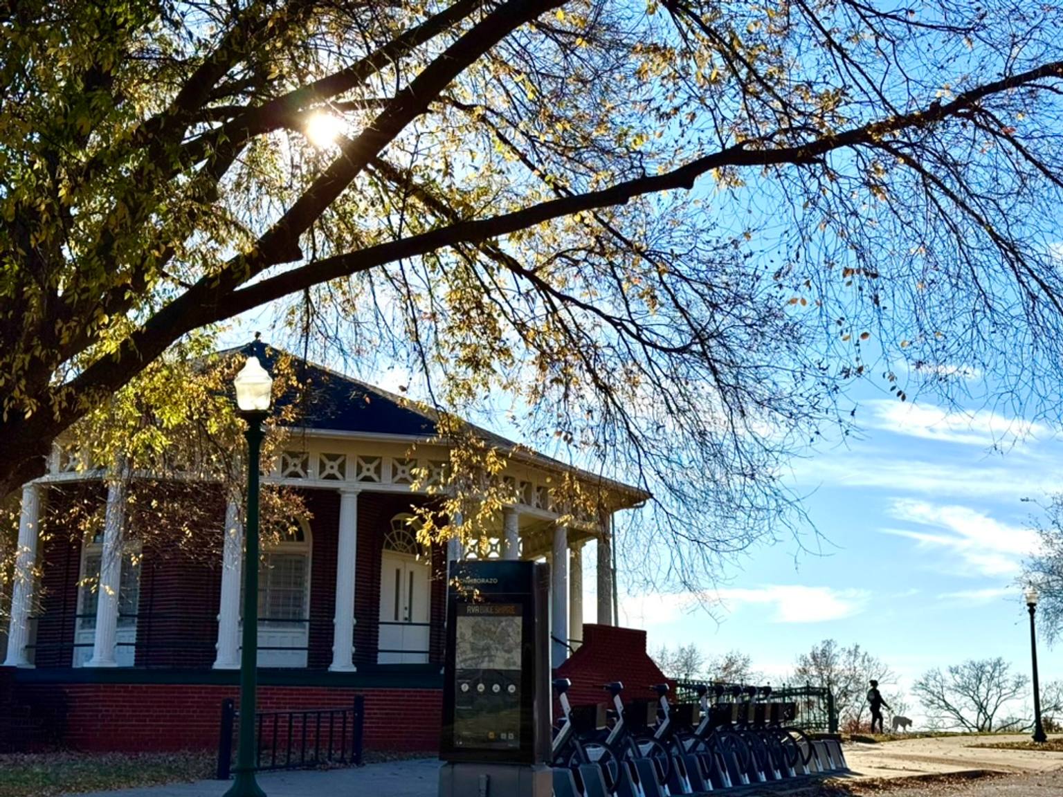 The historic Chimborazo Park Round House with white columns and a covered porch sits beneath a large willow tree with golden autumn foliage. A street lamp and informational marker are visible in front of the structure, with rows of chairs arranged on the grounds below.