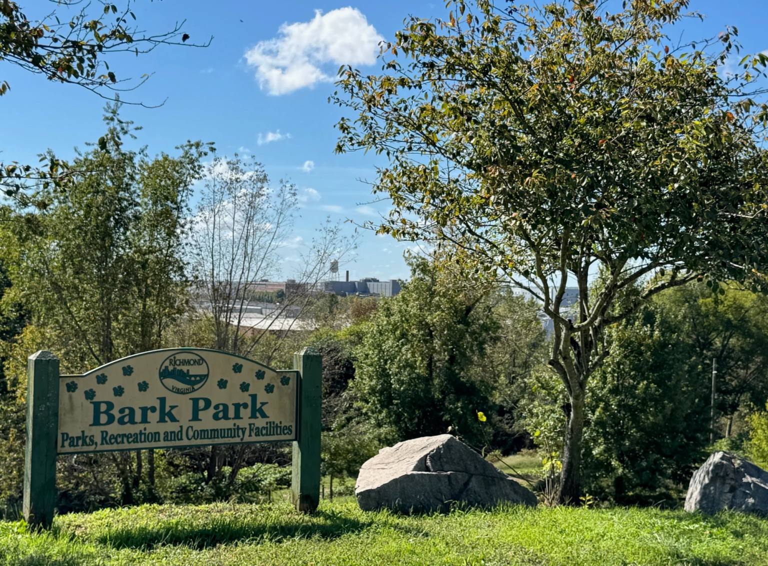 The Bark Park sign from Richmond's Parks, Recreation and Community Facilities department is displayed among trees and grass, with the Richmond industrial skyline and James River visible in the background.