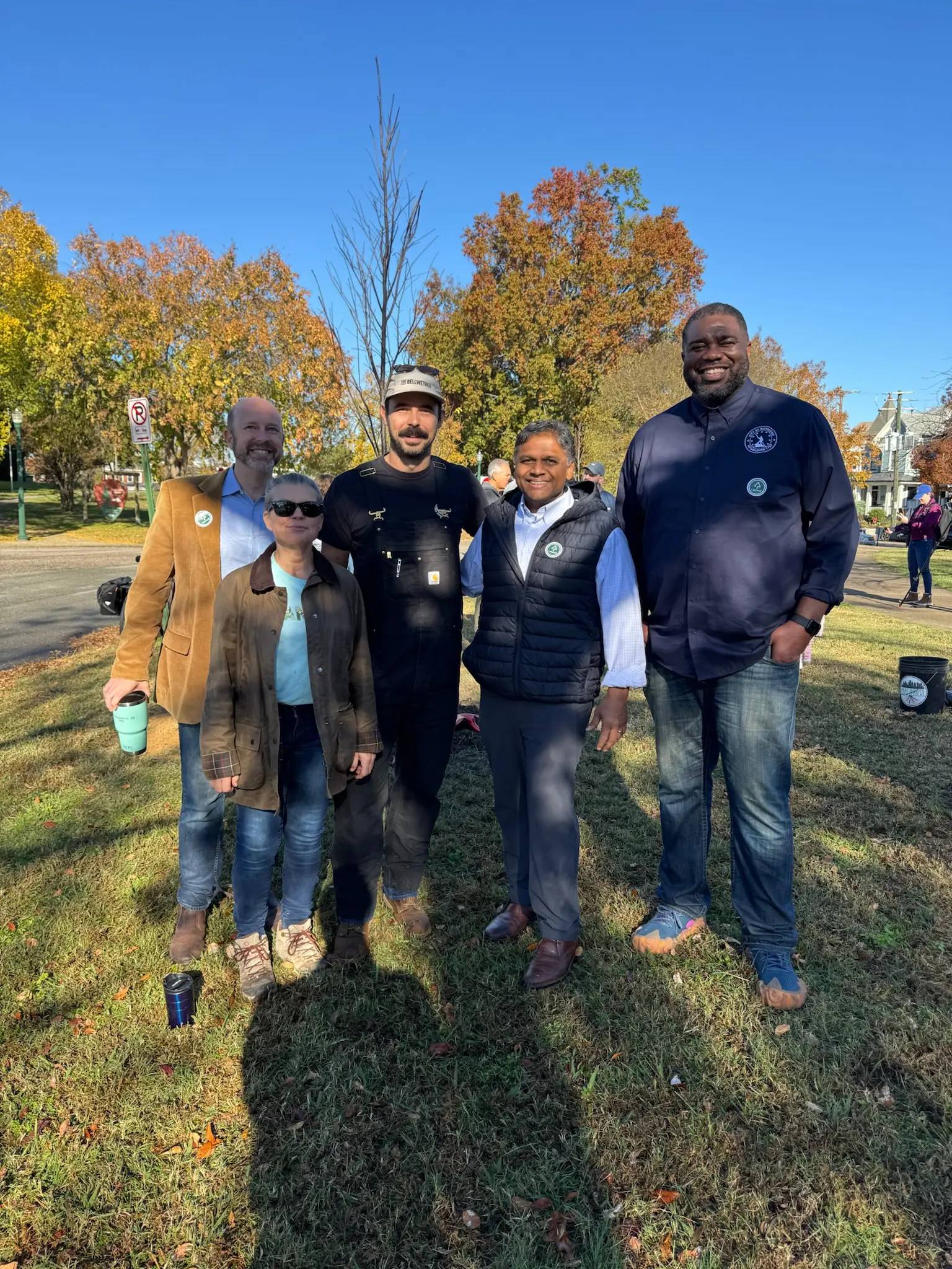Mayor Avula, city officials, and Conservancy board members pose together on the grass at Chimborazo Park, with autumn-colored trees in the background under a clear blue sky.