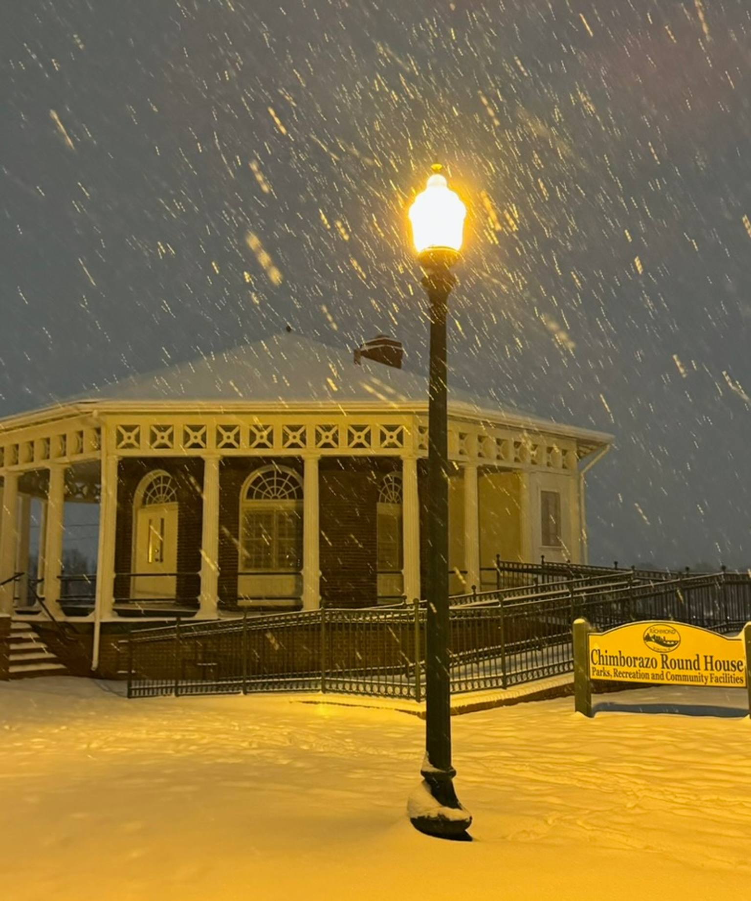 The historic Chimborazo Round House illuminated by a streetlamp during heavy snowfall at night, with snow accumulating on the yellow wooden deck and architectural details of the building visible in the background.