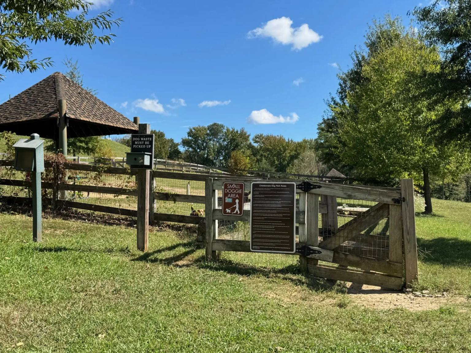 Fenced dog park entrance at Chimborazo Park with wooden gates, a covered pavilion, small dog area signage, and waste disposal station on a sunny day.