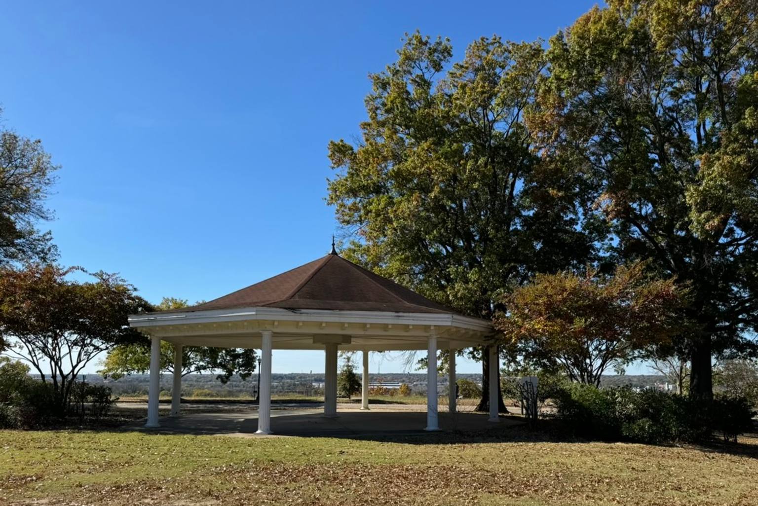 A white columned gazebo with a dark brown peaked roof stands in a grassy area surrounded by mature trees at Chimborazo Park, with scenic views of the surrounding landscape visible beyond the structure.