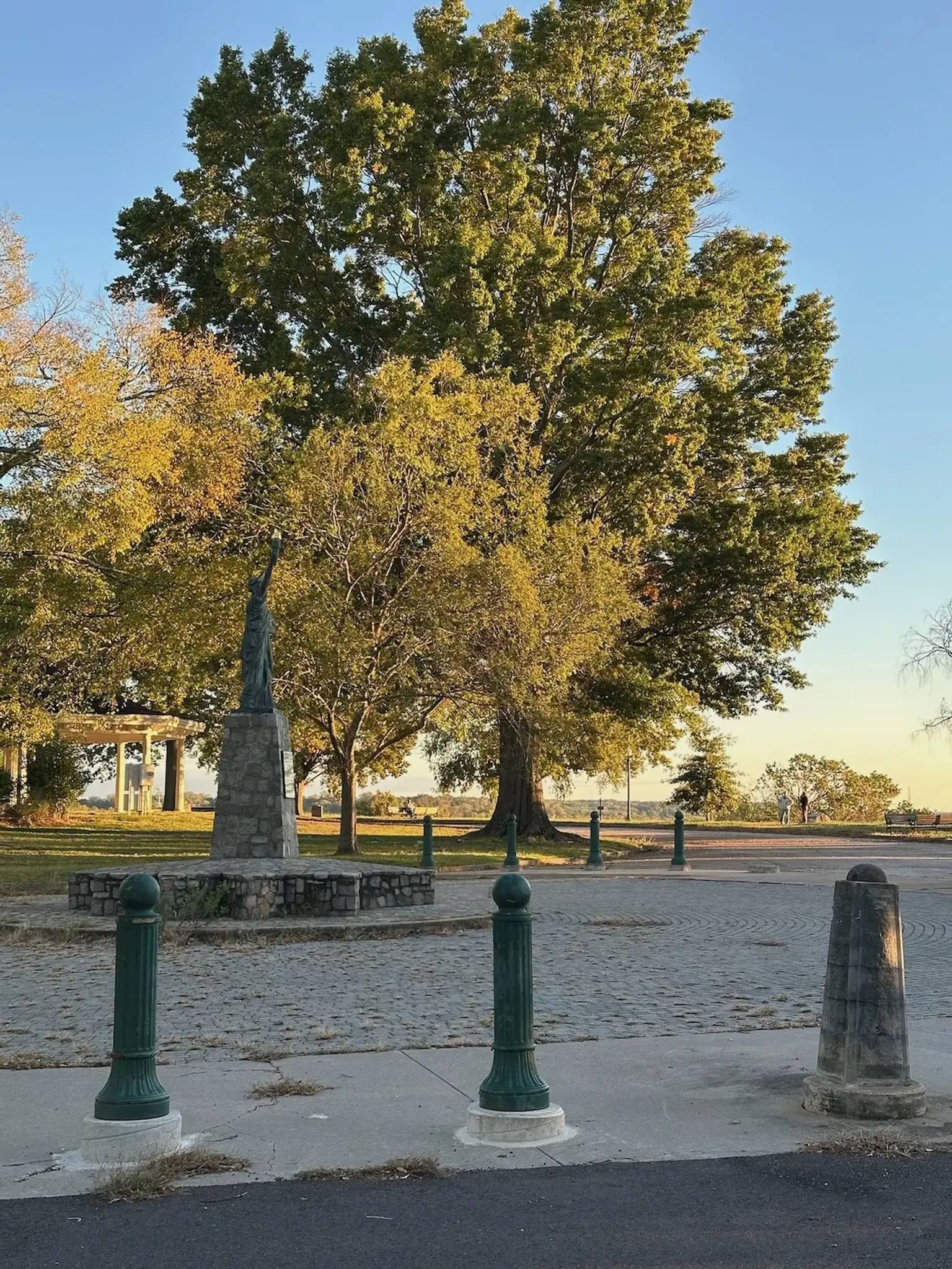 A scenic view of Chimborazo Park featuring three green bollards in the foreground on a paved area, with a stone monument and massive old oak trees displaying autumn foliage in the background, and a gazebo visible to the left.