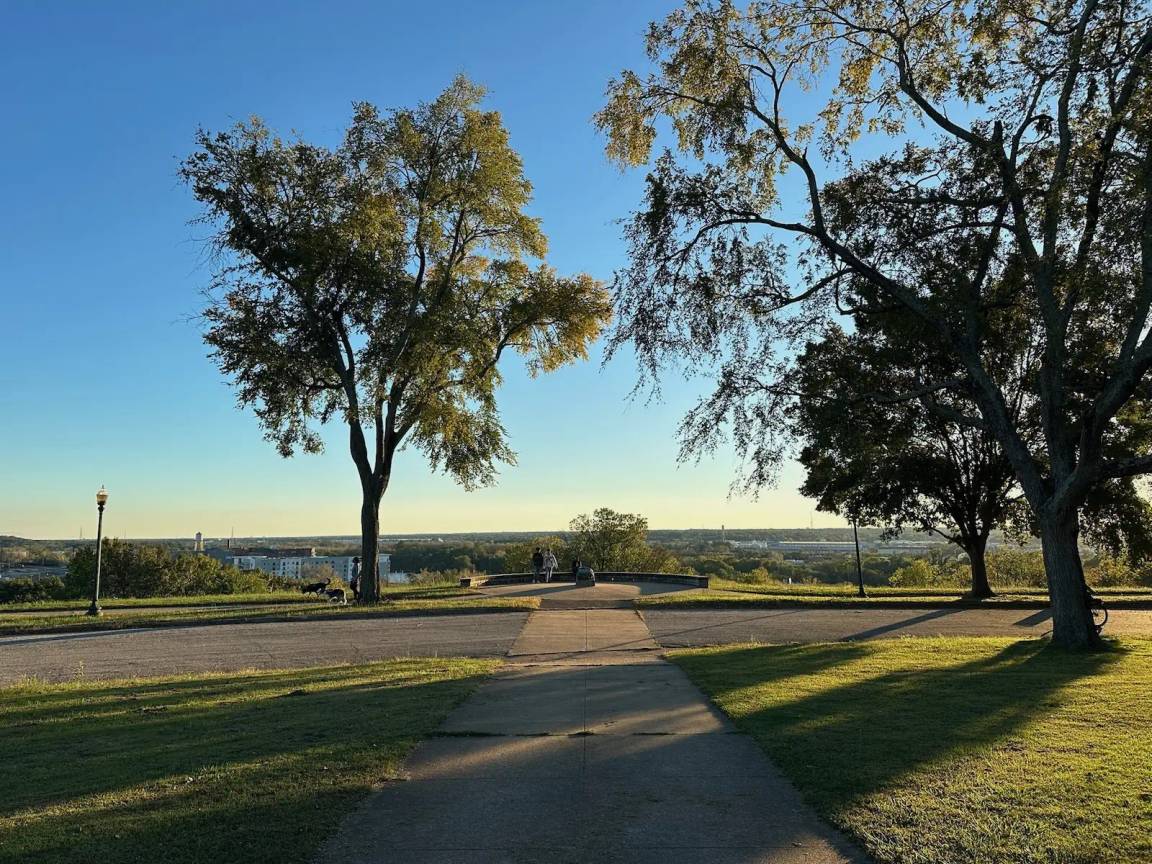 A tree-lined pathway in Chimborazo Park leading toward an overlook with a clear view of the James River and Richmond's skyline in the distance under a blue sky.