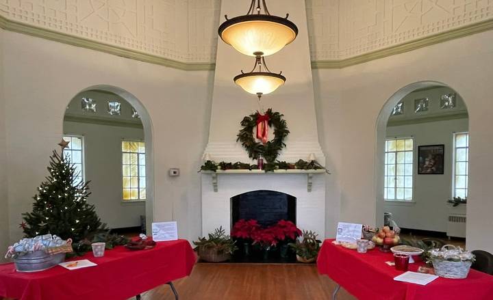 The Chimborazo Park Round House room decorated for the holiday season featuring a fireplace with red poinsettias, a wreath with red ribbon above the mantel, a decorated Christmas tree on the left, and two tables draped in red tablecloths with refreshments and displays.