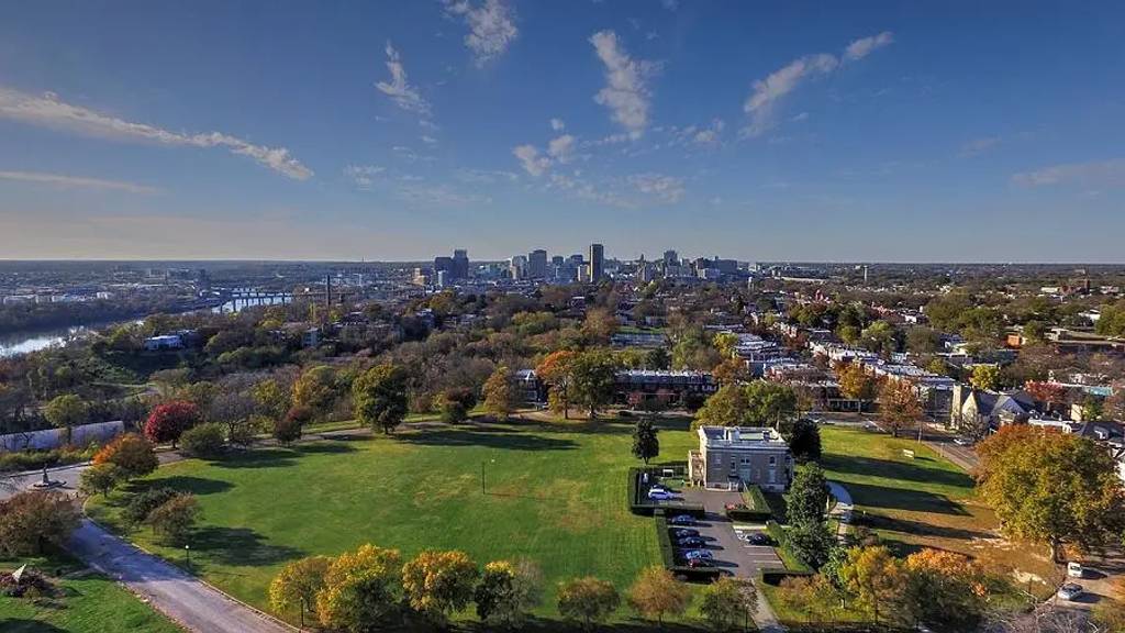 Aerial view of Chimborazo Park showing expansive green lawns and autumn foliage, with a modern building and parking area in the foreground, residential neighborhoods surrounding the park, and Richmond's downtown skyline visible across the James River in the distance.