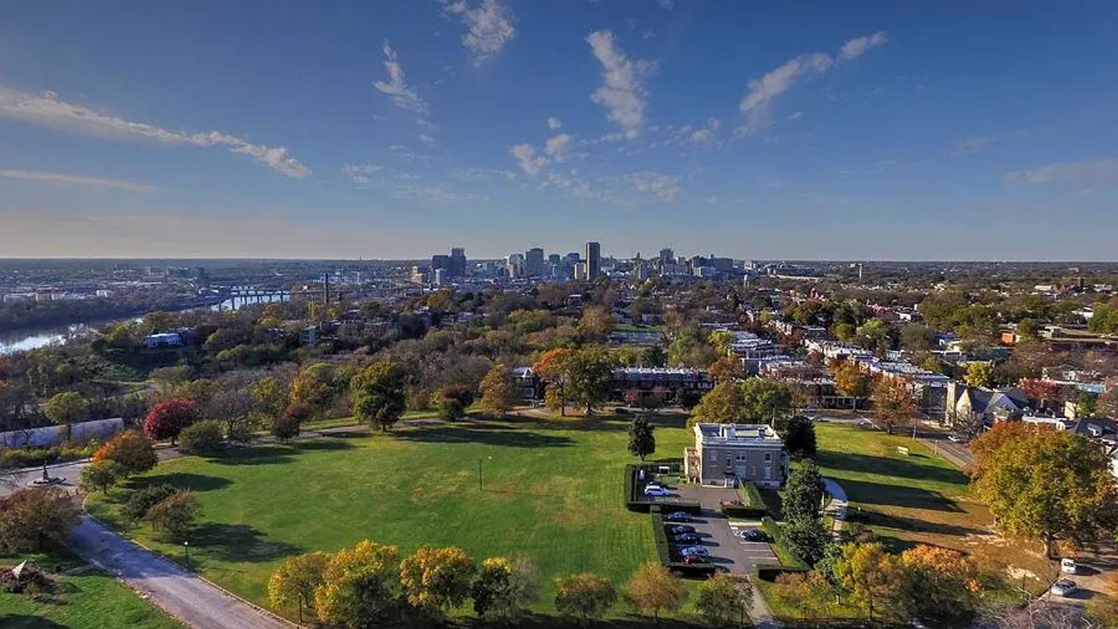 Aerial view of Chimborazo Park showing expansive green lawns and autumn foliage, with a modern building and parking area in the foreground, residential neighborhoods surrounding the park, and Richmond's downtown skyline visible across the James River in the distance.