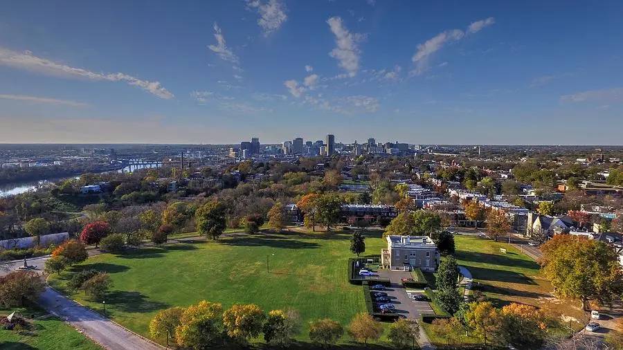 Aerial view of Chimborazo Park showing expansive green lawns and autumn foliage, with a modern building and parking area in the foreground, residential neighborhoods surrounding the park, and Richmond's downtown skyline visible across the James River in the distance.