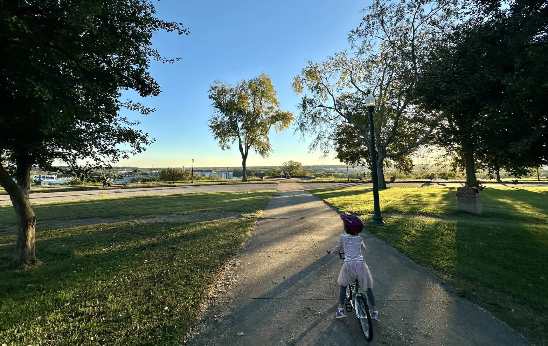 A person rides a bicycle on a paved path through Chimborazo Park, with large shade trees lining both sides of the path, open grassy areas, and a scenic view of the James River and Richmond's cityscape in the distance under a clear blue sky.