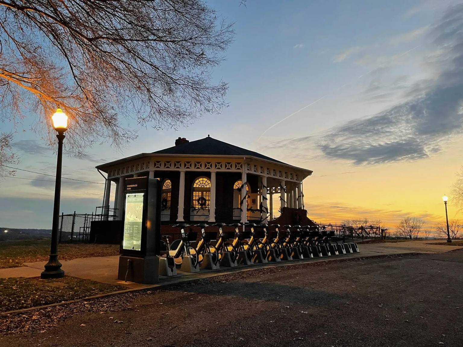 The historic Roundhouse structure in Chimborazo Park is illuminated at dusk, featuring a distinctive black octagonal pavilion with ornate railings, topped with a pyramid roof. A row of yellow bicycles from a bike-sharing program lines the front of the building, with a lit street lamp in the foreground and bare trees silhouetted against a golden sunset sky.