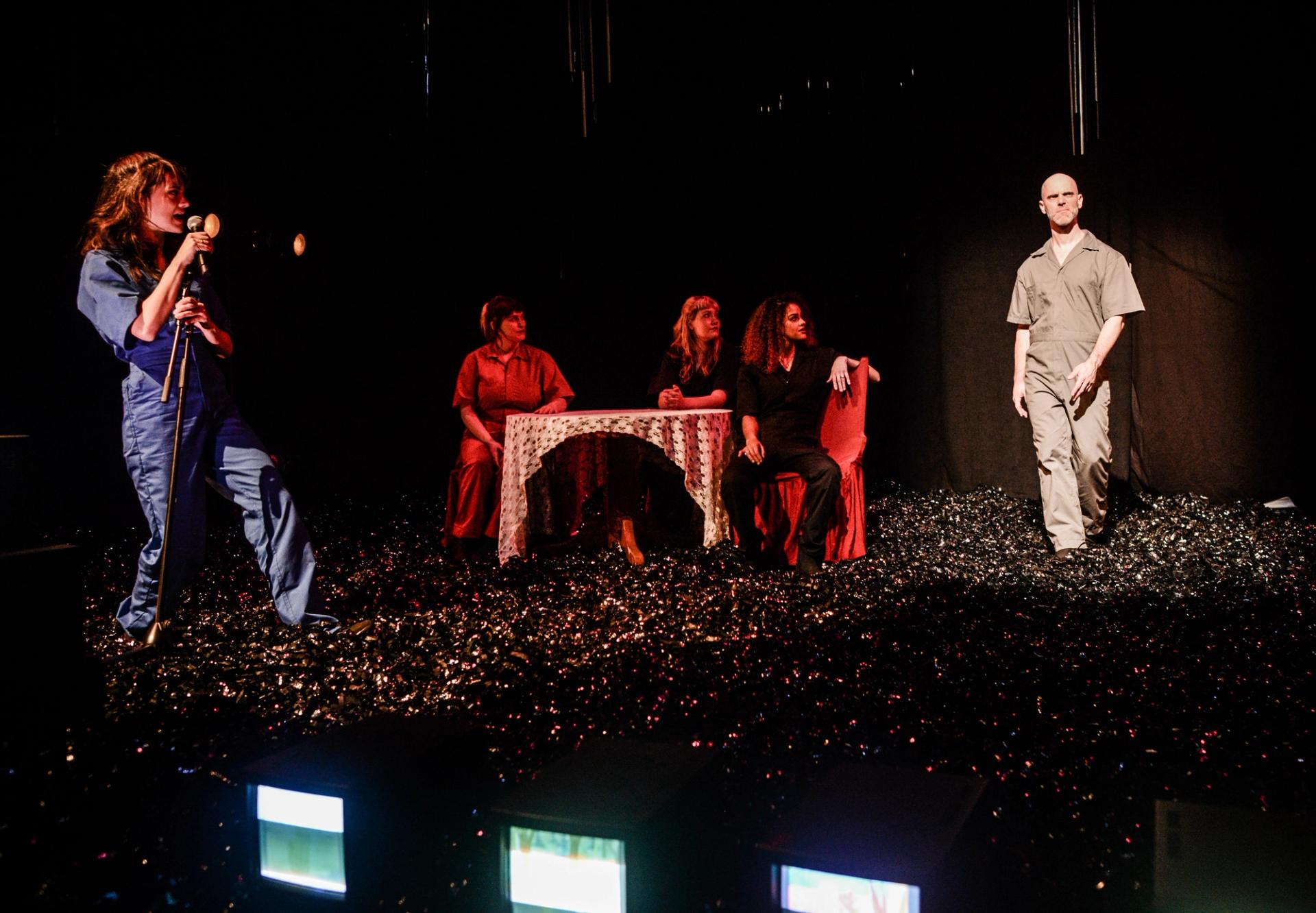 A woman speaks into a microphone as a man walks out into a spotlight with a look on his face as if he smells something strange.