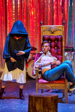 A robed women looms ominously next to a man on a throne.