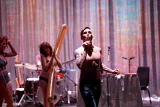 A shirtless man blows bubbles.